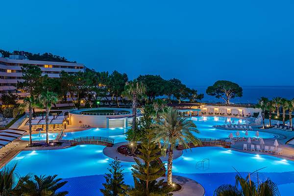 Nachtansicht des Rixos Premium Tekirova Pools mit Hotelgebäude und Meer im Hintergrund.