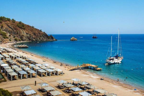 Luftaufnahme des Rixos Premium Tekirova Strandes mit vielen Sonnenliegen und Segelbooten im Wasser.