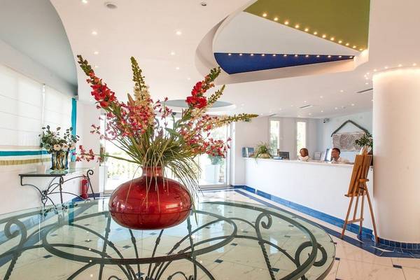 Empfangsbereich des Belvedere Luxury Suites Hotels mit rotem Blumenvase auf Glastisch und zwei Personen am Empfangstresen.