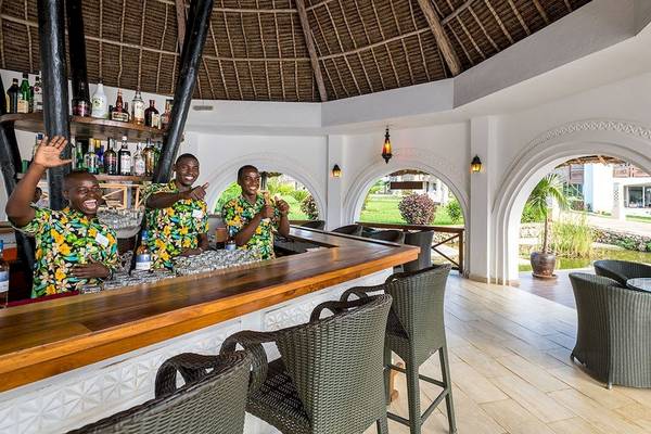 Drei freundliche Barkeeper im Royal Zanzibar Beach Resort, vor einer Bar mit vielen Gläsern und Flaschen.