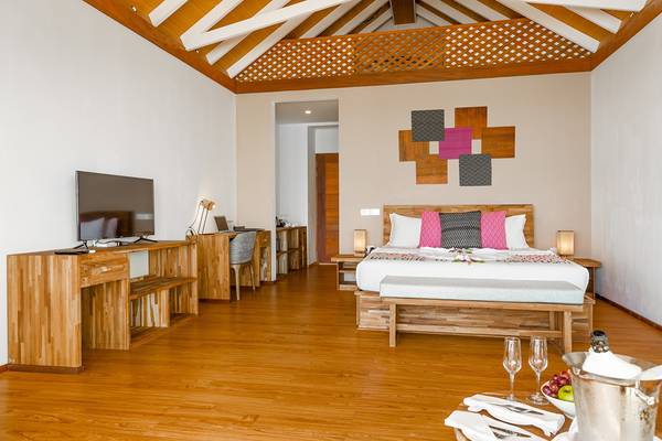Innenansicht einer Suite im Kudafushi Resort & Spa mit einem großen Bett, Fernseher, Schreibtisch und Dekor.