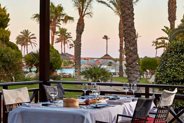 Terrasse mit Tisch und Blick auf Palmen und eine Poollandschaft im Atlantica Aegean Blue Resort.