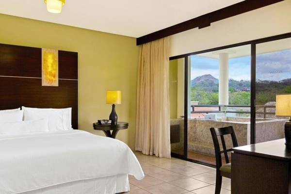 Innenansicht eines Hotelzimmers im Westin Reserva Conchal, mit Bett, Tisch, Lampen und Balkon.