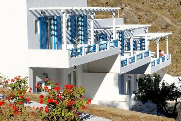Außenansicht des Rhenia Mykonos Hotel & Bungalows mit weißen Wänden, blauen Fensterläden und Balkonen, umgeben von roten Blumen und Pflanzen.