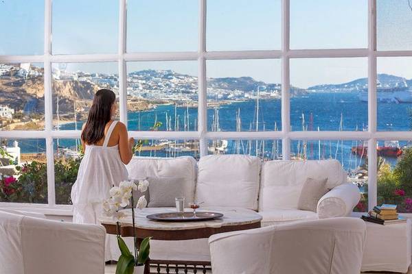 Frau im weißen Kleid mit Blick aus dem Fenster des Rhenia Mykonos Hotels auf den Hafen und die Bucht.