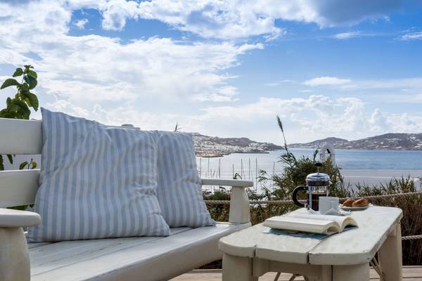Terrasse im Rhenia Mykonos Hotel mit Aussicht auf das Meer, einem weißen Sofa mit blauen Kissen, einem Tisch mit einem Buch, Kaffee und Brot.