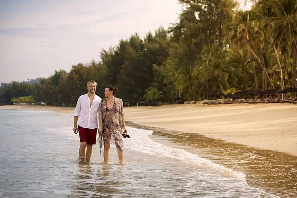 Ein Paar steht am Strand von Robinson Khao Lak, umgeben von Bäumen und Sonnenliegen.