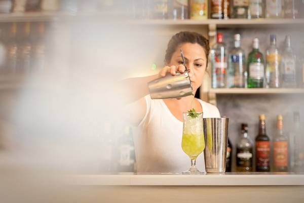 Frau mischt einen Cocktail an einer Bar mit vielen Flaschen im Hintergrund.