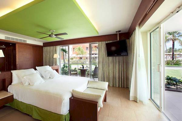 Hotelzimmer mit großem Bett, grüner Decke, Fernseher, Glastüren und Blick auf den Garten.