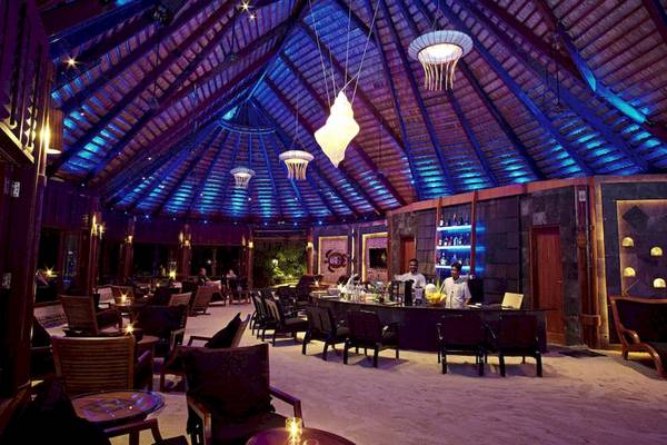 Innenansicht der Bar im Komandoo Island Resort & Spa mit blauer Beleuchtung und runden Tischen.