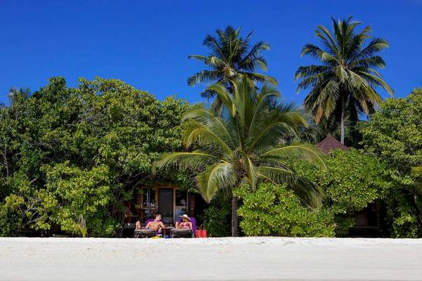 Zwei Personen entspannen auf dem Strand vor dem Komandoo Island Resort & Spa, umgeben von Palmen.