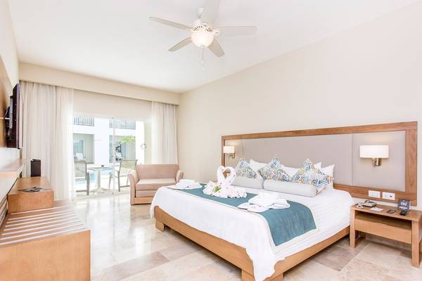 Innenansicht eines Hotelzimmers im Sunscape Coco Punta Cana mit Bett, Wohnzimmermöbeln und Swimmingpool im Hintergrund.