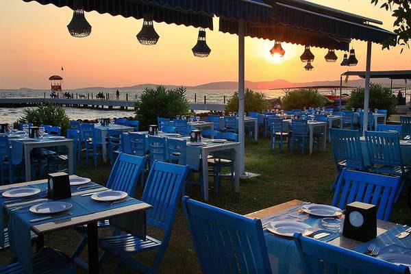 Außenbereich des Club Yali Hotels & Resort bei Sonnenuntergang mit blauen Stühlen und Tischen am Meer.