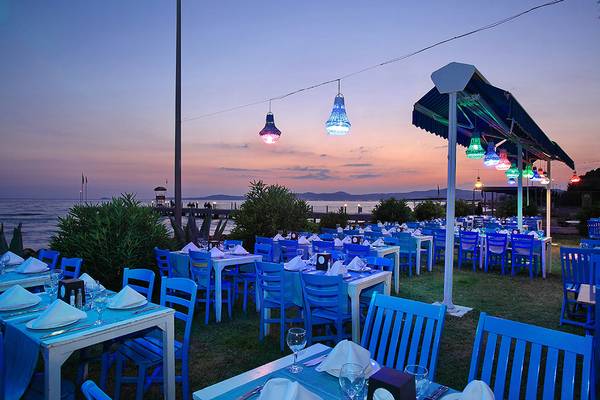 Außenbereich des Club Yali Hotels & Resort bei Sonnenuntergang mit blauen Stühlen und Tischen.