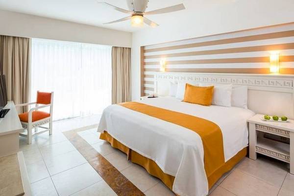 Modernes Hotelzimmer im BlueBay Grand Esmeralda mit weißem Bett, orangen Kissen, Fernseher, Stuhl und Deckenventilator.