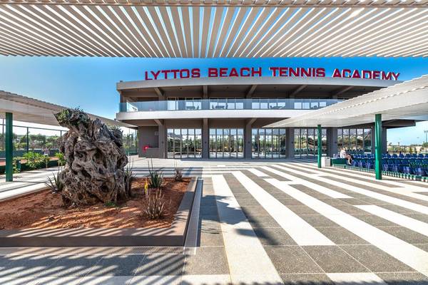 Außenansicht des Lyttos Beach Tennis Academy mit einer großen Baumskulptur und einem blauen Himmel.