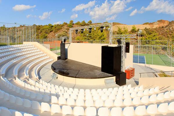 Ein Freilufttheater mit Sitzreihen und einer Bühne, umgeben von einem Zaun und einem Tennisplatz im Hintergrund.