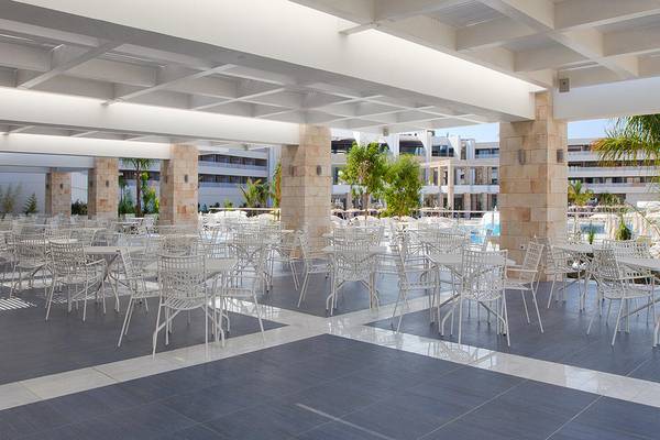 Lounge-Bereich des Princess Andriana Resort & Spa mit weißen Tischen und Stühlen unter einer überdachten Terrasse.
