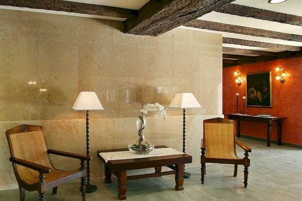 Hotellobby des Occidental Playa de Palma mit beigefarbenen Wänden, Holzbalkendecke, zwei Sesseln, einem Tisch mit Blumenvase und zwei Lampen.