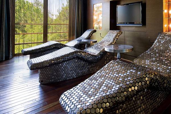 Loungebereich im Cornelia Diamond Golf Resort & Spa mit modernen, metallisch glänzenden Liegestühlen und Fernseher.