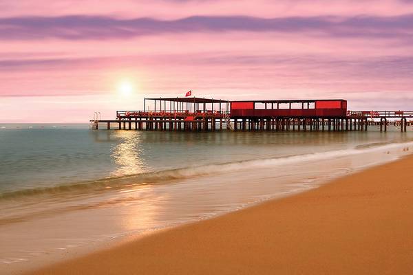 Sonnenuntergang am Pier des Cornelia Diamond Golf Resort & Spa mit rotem Gebäude und Flagge.