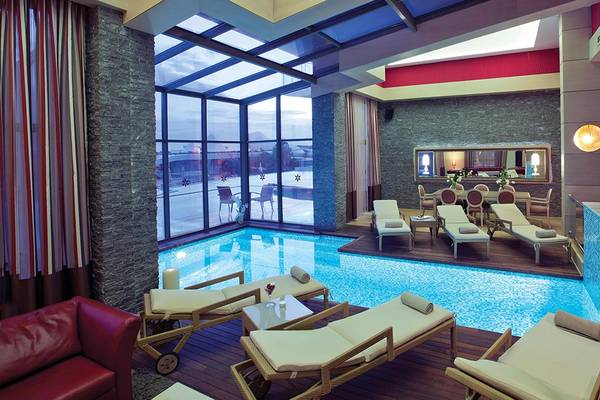 Innenansicht des Cornelia Diamond Golf Resort & Spa mit Pool, Liegestühlen und Glaswänden.