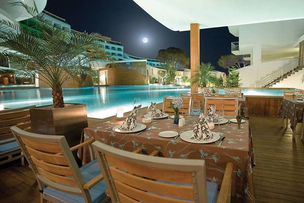 Außenbereich des Cornelia Diamond Golf Resort & Spa bei Nacht mit gedecktem Tisch und Swimmingpool im Hintergrund.