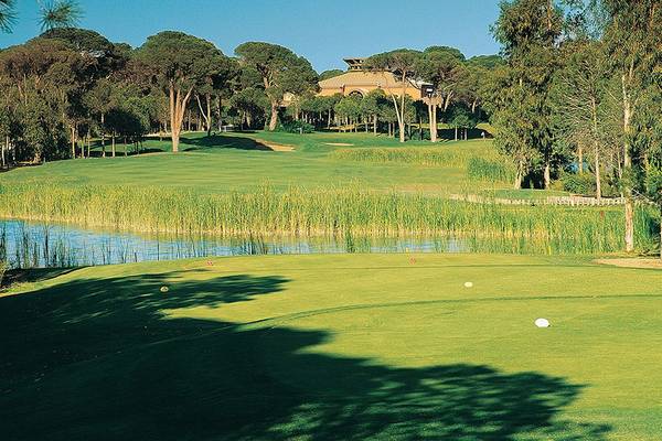 Golfplatz des Cornelia Diamond Golf Resort & Spa mit einem Gebäude im Hintergrund und einem See vor der Anlage.