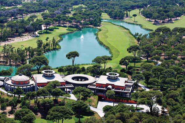 Luftaufnahme des Cornelia Diamond Golf Resort & Spa mit einem Golfplatz, einem See und umliegenden Bäumen.
