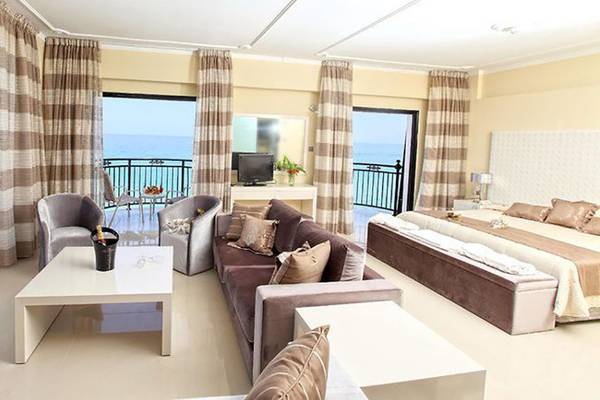 Innenansicht eines Hotelzimmers im Constantinos the Great Beach Hotel mit Meerblick, einem Doppelbett, einem Sofa, einem Tisch und einem Fernseher auf einem Tisch.