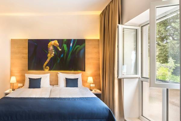 Doppelzimmer im Remisens Hotel Epidaurus mit Meerblick, modernes Design, Meerpferd auf dem Wandbild, zwei Lampen auf Nachttischen, offenes Fenster.