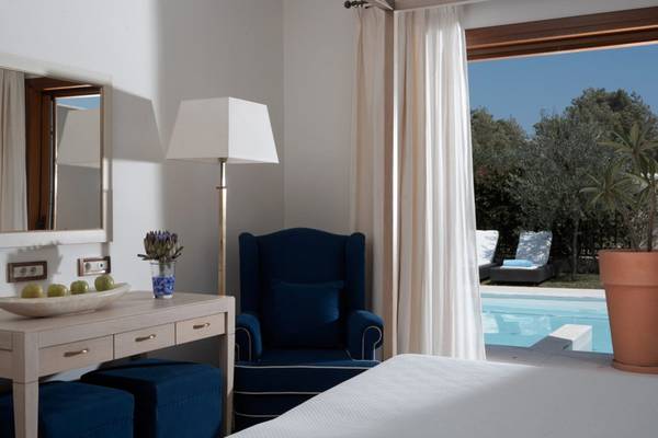 Innenansicht eines Zimmers im Lindian Village Beach Resort auf Rhodos mit einem Bett, einem blauen Sessel und einer Aussicht auf einen Pool.