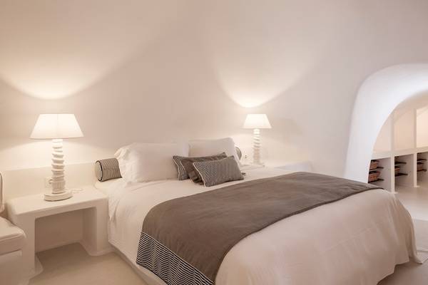 Ein geräumiges Hotelzimmer im Katikies Chromata Santorini mit weißen Wänden, zwei Lampen, einem Bett und einem weißen Nachttisch.