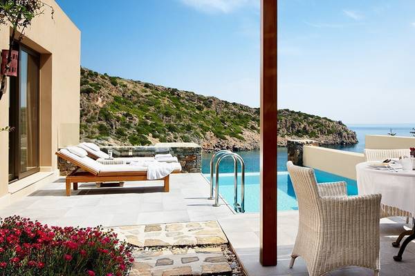 Außenbereich des Daios Cove Luxus Resorts mit Swimmingpool und Meerblick, Liegestühlen und Esstisch.
