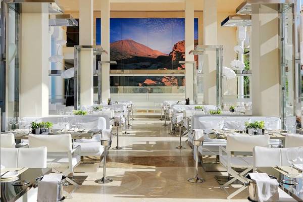 Innenbereich des Gran Melia Palacio de Isora Restaurants mit vielen weißen Tischen und Stühlen und einer großen Glaswand.