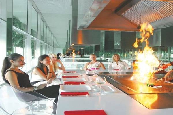 Eine Gruppe von Frauen sitzt am Tisch in einem modernen Restaurant im Adam & Eve Hotel, während ein Koch ein Gericht auf einem offenen Grill vorbereitet.