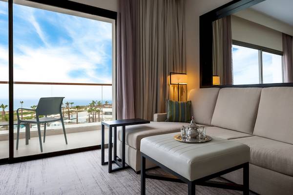 Ein modernes Hotelzimmer mit einem großen Balkon, der Meerblick bietet. Im Zimmer steht eine große Couch, ein Couchtisch und ein kleiner Tisch mit einer Lampe.