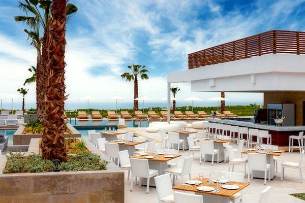 Außen-Essbereich des Hyatt Place Taghazout Bay Hotels mit weißen Tischen und Stühlen, Swimmingpool und Palmen