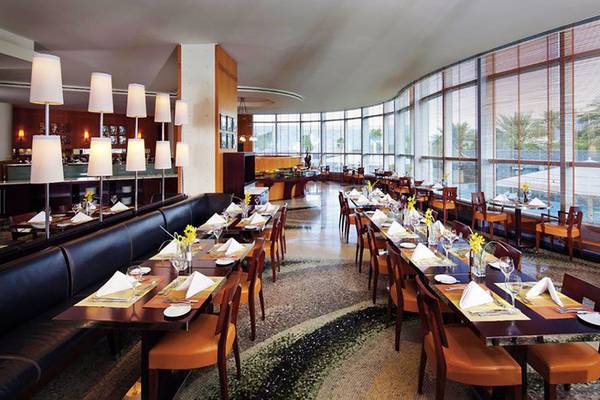 Innenansicht des Restaurants im Jumeirah Emirates Towers Hotel mit runden Tischen und Stühlen, große Fenster und Außenpool im Hintergrund.
