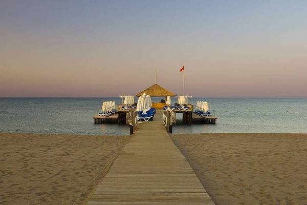 Ein Holzsteg führt zur Strandbar des Blue Waters Club, mit Sonnenliegen und Sonnenschirmen.