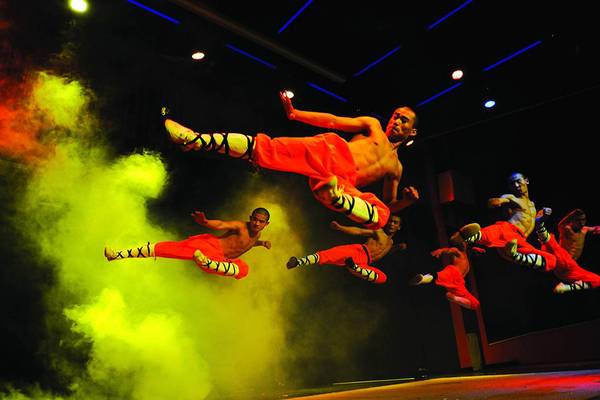 Mehrere Männer in roten Kostümen springen in der Luft, während sie eine Choreografie aufführen, während Raucheffekte um sie herum sind.