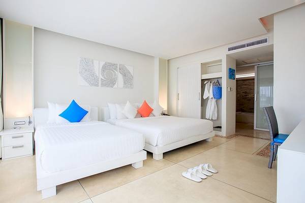 Innenansicht eines Doppelzimmers im Samui Resotel Beach Resort mit zwei weißen Betten, blauen und orangefarbenen Kissen, weißen Nachttischen und einem offenen Kleiderschrank.