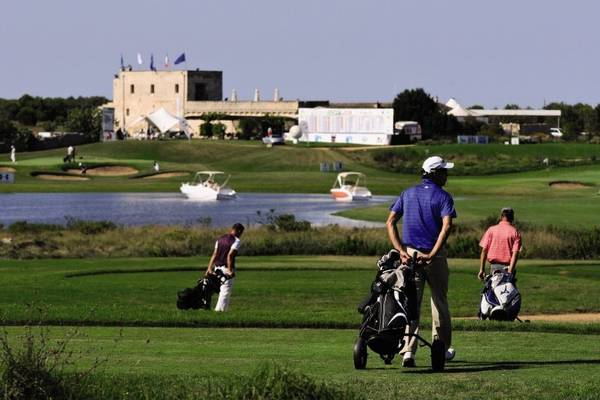 Golfer auf dem Golfplatz des MIRA Acaya Golf Resort & SPA mit Gebäude und Wasser im Hintergrund.