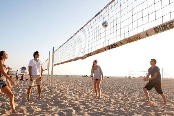 Vier Personen spielen Volleyball am Strand mit einem Netz, das Duke und Sandy & Duke beschriftet ist.