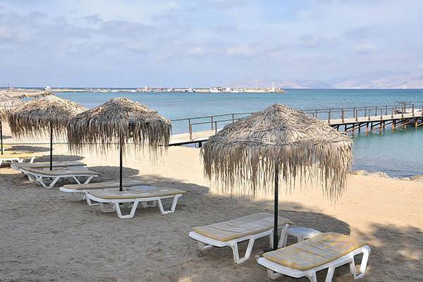 Strandszene mit mehreren Sonnenliegen, großen Palmenhügelschirmen und einem Steg im Hintergrund.