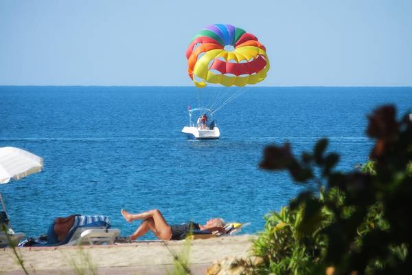 Ein Parasailing-Erlebnis am Strand des Riviera Hotels & Spa mit einer bunten Drachenparachute und zwei Personen, die auf Liegestühlen entspannen.