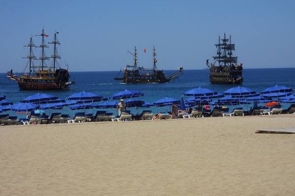 Sonnenbadende Gäste auf einem Strand mit Segelschiffen im Hintergrund.
