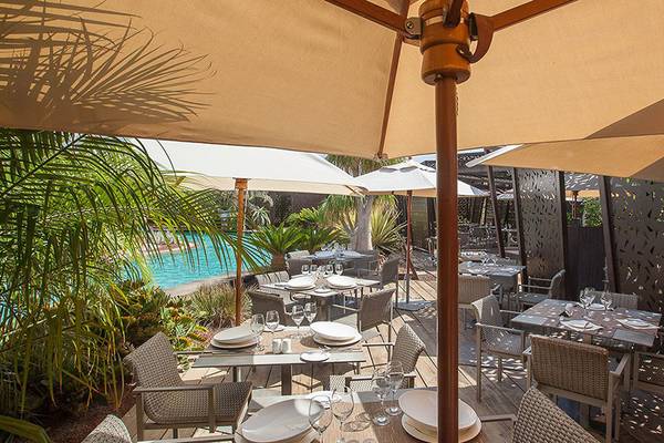 Außenbereich des Lopesan Baobab Resorts mit Tischen und Stühlen unter Sonnenschirmen und Blick auf den Pool.