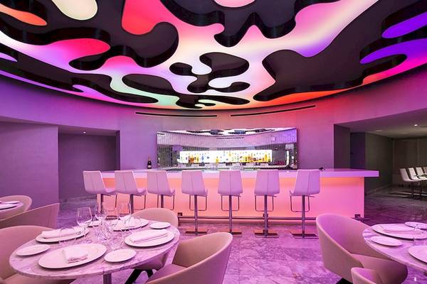 Hotel-Bar-Lounge-Bereich mit Tisch, Barhockern und violett-pinker Beleuchtung.