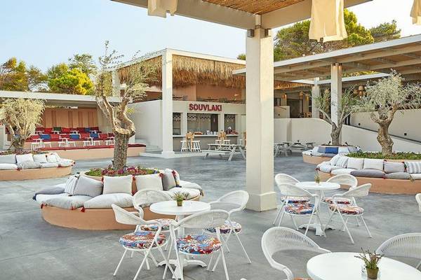Außenbereich des Grecotel Olympia Oasis Aqua Park mit Tischen, Stühlen und einem großen Baum.